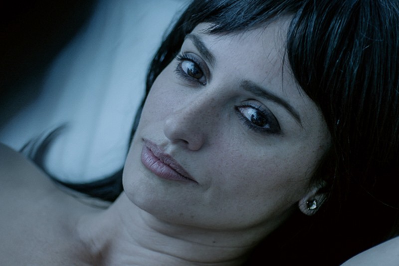 Kobieta o ciemnych włosach i lekkim makijażu leży na boku, patrząc łagodnie w kamerę ze spokojnym, zamyślonym wyrazem twarzy. Nosi mały kolczyk i odpoczywa na białej poduszce.