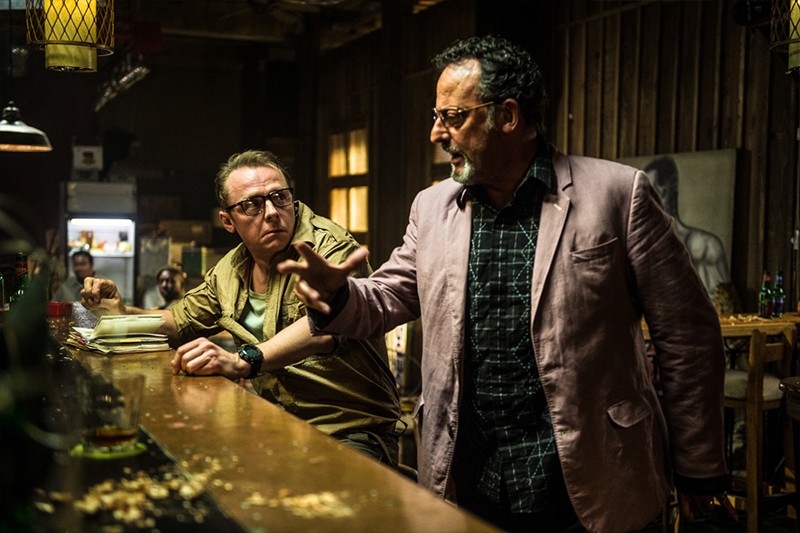 Dwóch mężczyzn intensywnie rozmawia przy zagraconym barze w słabo oświetlonym pomieszczeniu. Jeden z nich nosi okulary i brązową marynarkę, siedzi, podczas gdy drugi, stojący, gestykuluje stanowczo w jasnej marynarce i kraciastej koszuli.