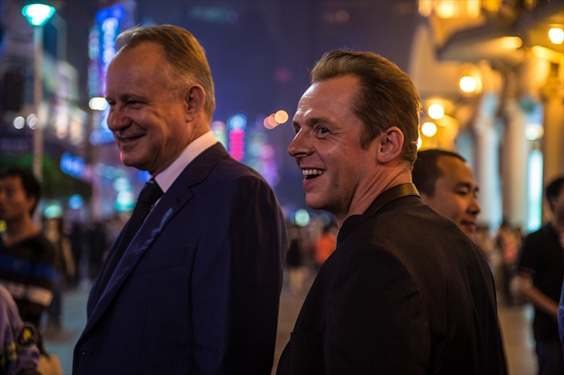 Dwóch mężczyzn w garniturach uśmiecha się, stojąc nocą na jasno oświetlonej ulicy miasta, otoczonych przez innych ludzi. Neony i latarnie uliczne oświetlają tętniące życiem miejskie tło.