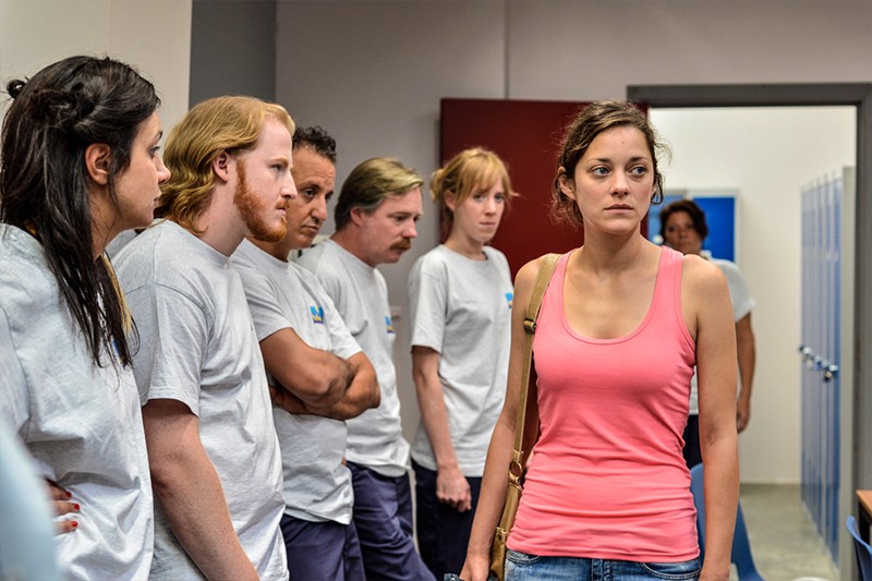 Kobieta w różowym topie stoi naprzeciwko grupy ludzi w szarych koszulkach, którzy patrzą na nią z poważnymi minami na korytarzu, w tle widać szafki i otwarte drzwi.