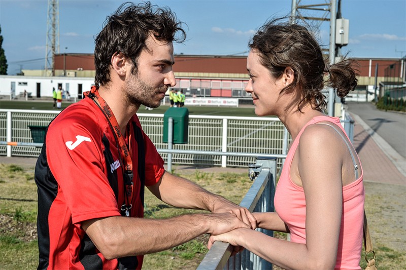 Mężczyzna w czerwonej koszulce sportowej trzyma za rękę kobietę w różowym topie po drugiej stronie metalowego płotu. Oboje uważnie się sobie przyglądają w słoneczny dzień, niedaleko boiska sportowego.