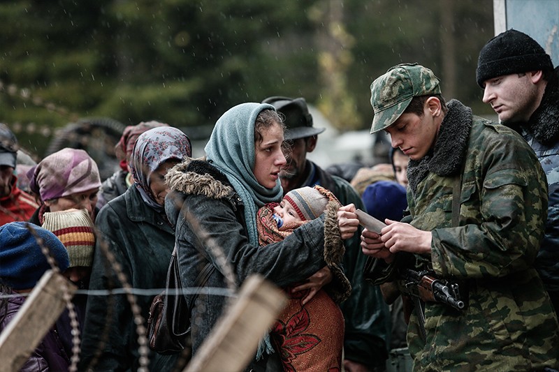Grupa ludzi w znoszonych ubraniach stoi na zewnątrz za drutem kolczastym, podczas gdy żołnierz w kamuflażu sprawdza dokumenty kobiety. Kobieta trzyma dziecko owinięte w koc; inni czekają w kolejce pod szarym niebem.