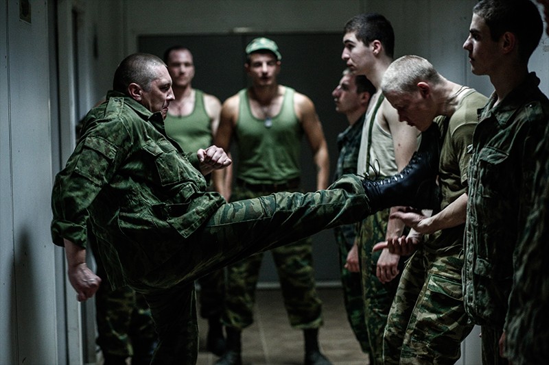 Mężczyzna w mundurze wojskowym kopie innego mężczyznę stojącego w szeregu młodych mężczyzn, wszyscy ubrani w stroje wojskowe, w słabo oświetlonym korytarzu. Inni patrzą, niektórzy stoją na baczność.