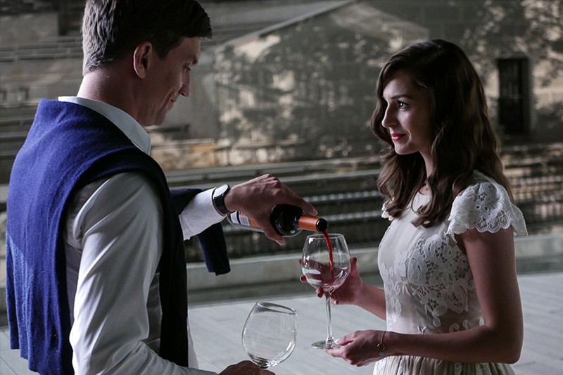 Mężczyzna nalewa kobiecie czerwone wino do kieliszka, gdy stoi na zewnątrz. Mężczyzna ma na sobie niebieski sweter na ramionach, a kobieta jest ubrana w białą koronkową sukienkę. Patrzą na siebie, ich wyrazy twarzy są spokojne i delikatne.