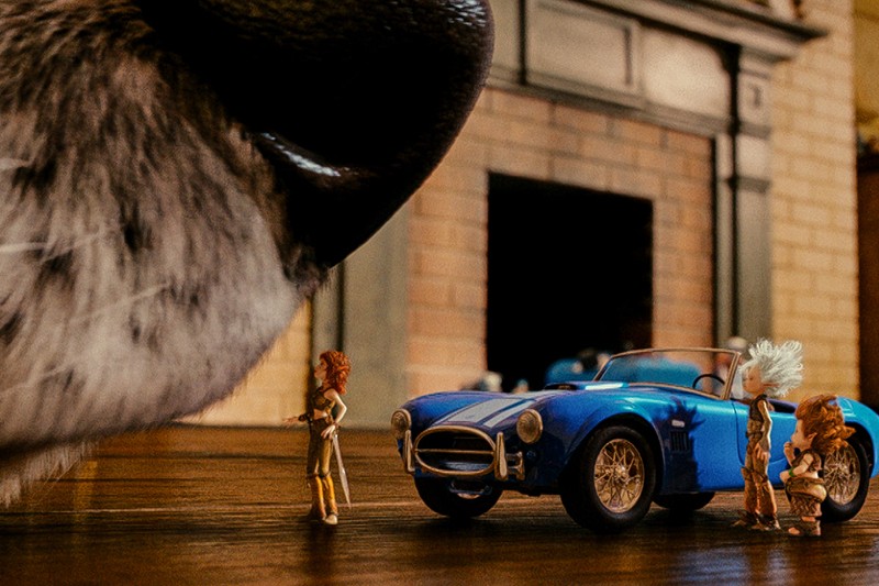 Nos dużego zwierzęcia unosi się nad trzema małymi figurkami i niebieskim samochodzikiem na drewnianej podłodze, w tle widać ceglany kominek.