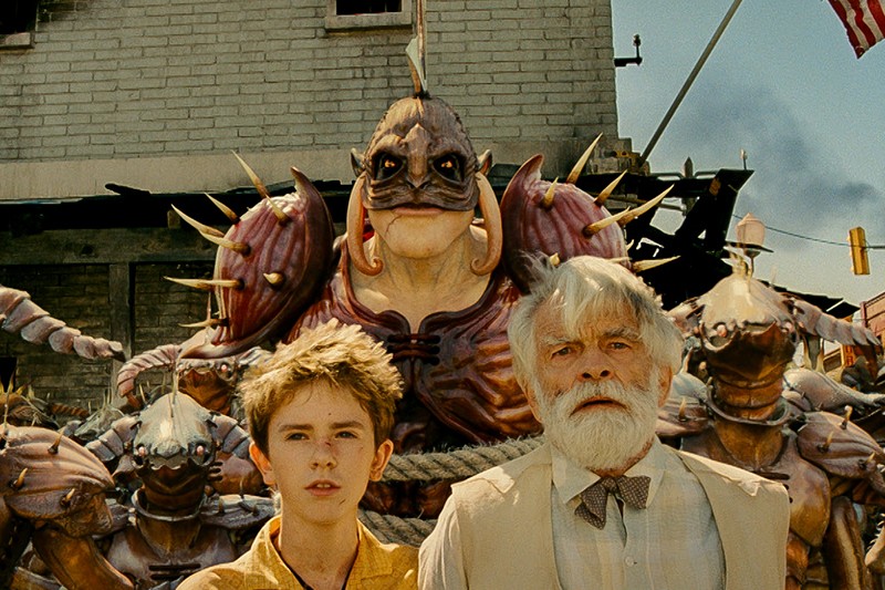 Młody chłopak i starszy mężczyzna z białymi włosami i brodą stoją na tle kolczastych, opancerzonych stworzeń przypominających ryby na miejskiej ulicy. W tle widoczna jest amerykańska flaga.