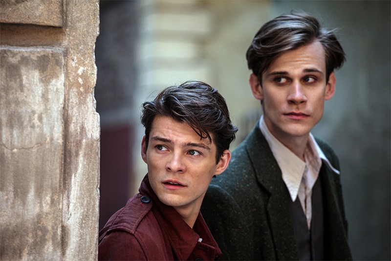 Dwóch młodych mężczyzn o poważnych minach stoi blisko siebie przy ścianie, patrząc w różnych kierunkach, jakby byli ostrożni lub zaniepokojeni. Jeden nosi czerwoną kurtkę, drugi zielony płaszcz i białą koszulę. Tło jest miejskie i stonowane.