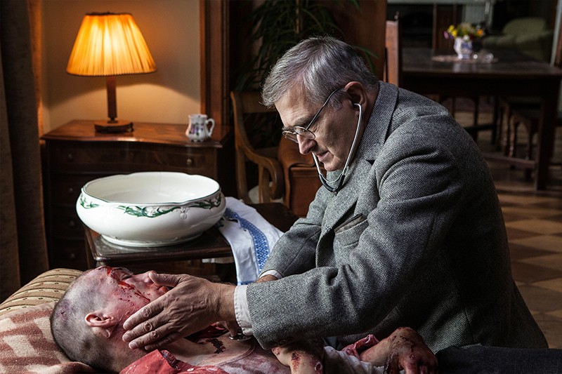 Starszy mężczyzna w okularach i szarym garniturze używa stetoskopu, aby zbadać ranną, zakrwawioną osobę leżącą na kanapie w słabo oświetlonym pokoju z lampą i meblami w tle.