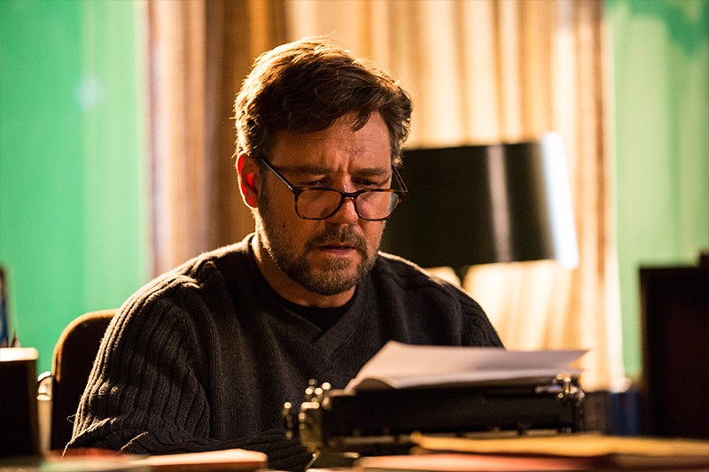 Mężczyzna w okularach i z brodą siedzi przy biurku, czyta dokumenty przed maszyną do pisania, w tle widać lampę i zasłony.