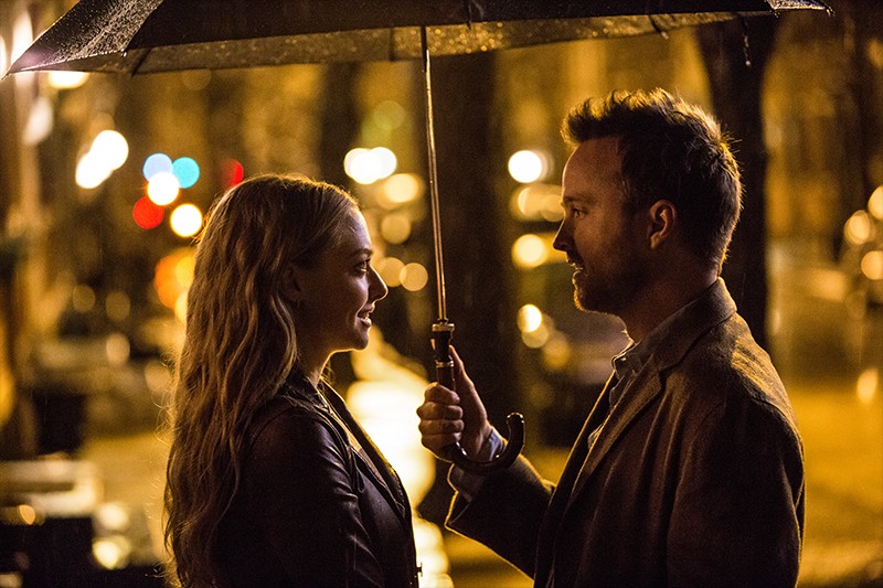 Mężczyzna i kobieta stoją naprzeciw siebie pod parasolem w deszczową noc, oświetleni ciepłym światłem latarni ulicznych. W tle widać rozmyte światła miasta i mokre ulice, tworząc romantyczną atmosferę.