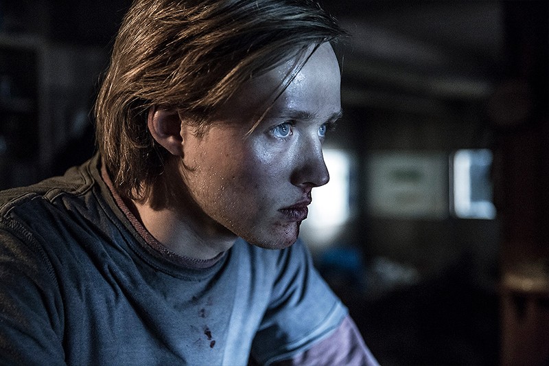 Młoda osoba z potarganymi włosami i posiniaczonymi, zakrwawionymi ustami intensywnie patrzy przed siebie w słabo oświetlonym pokoju, ubrana w szarą koszulę z widocznymi plamami krwi.