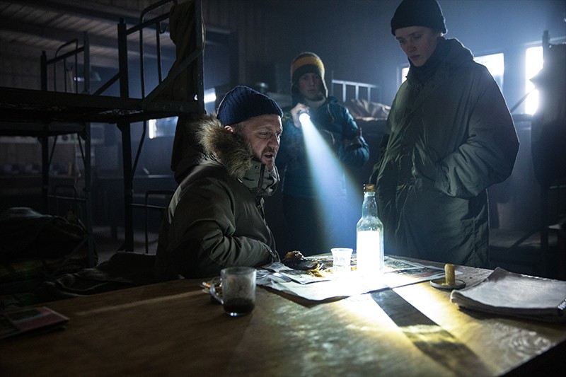 Trzy osoby w zimowych ubraniach stoją i siedzą przy stole w słabo oświetlonym pokoju z łóżkami piętrowymi. Snop światła pada na butelkę, szklankę i jedzenie, tworząc dramatyczne cienie. Atmosfera wydaje się napięta i ponura.