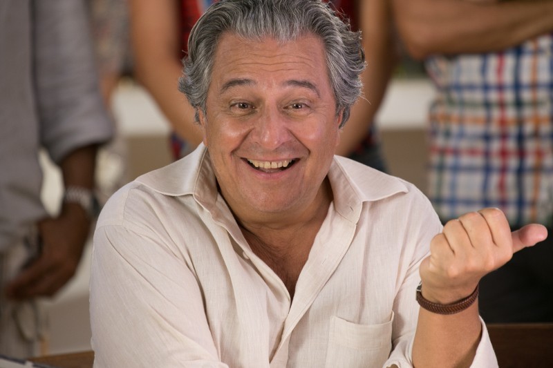 Mężczyzna w średnim wieku z siwymi włosami, ubrany w jasną koszulę, uśmiecha się ciepło i gestykuluje prawą ręką. Tło przedstawia ludzi stojących i rozmazanych, co sugeruje swobodną atmosferę towarzyską.