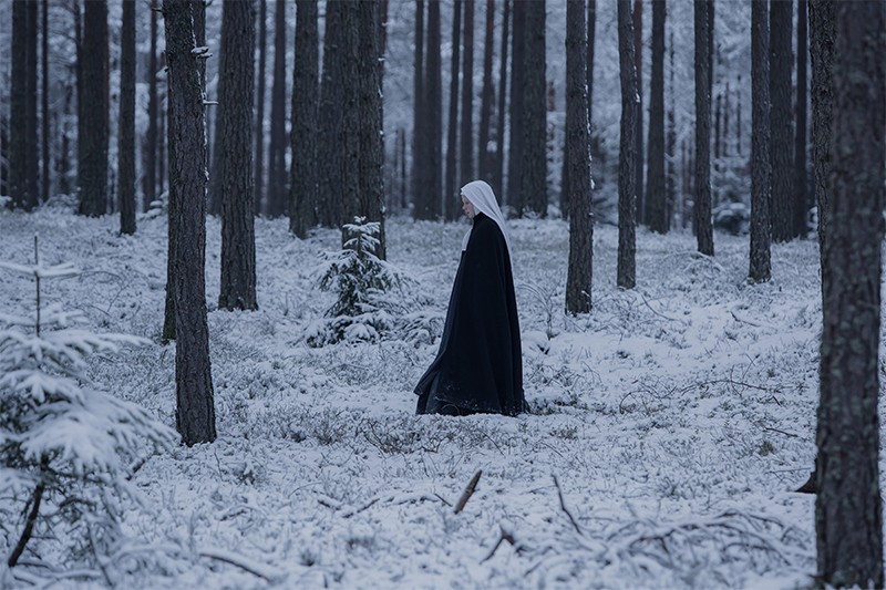 Osoba ubrana w czarny płaszcz i białe nakrycie głowy idzie samotnie przez zaśnieżony las, w otoczeniu wysokich drzew pokrytych śniegiem.