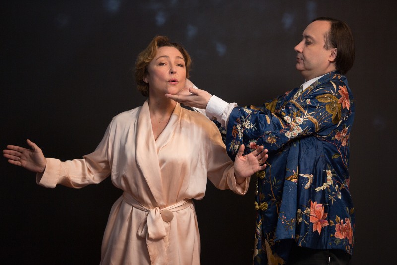 Kobieta w jasnoróżowej szacie śpiewa z wyciągniętymi przed siebie ramionami, podczas gdy mężczyzna w niebieskiej kwiecistej kurtce delikatnie podtrzymuje jej brodę, korygując jej postawę na ciemnym tle.