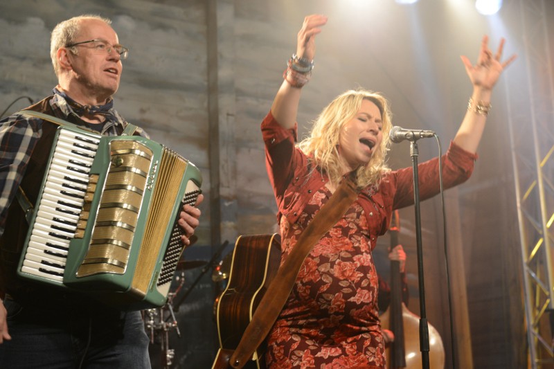 Mężczyzna gra na akordeonie, podczas gdy kobieta w wzorzystej sukience śpiewa namiętnie do mikrofonu na scenie, unosząc ramiona. W tle widoczne są światła sceniczne i instrumenty muzyczne.