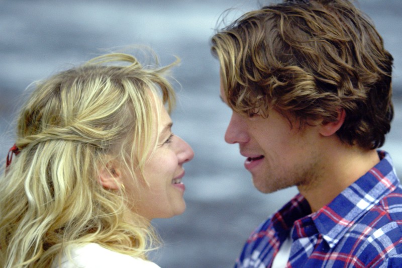 Blondynka i mężczyzna o falowanych brązowych włosach stoją blisko siebie, zwróceni ku sobie i uśmiechnięci, na tle rozmazanego, szarego tła.