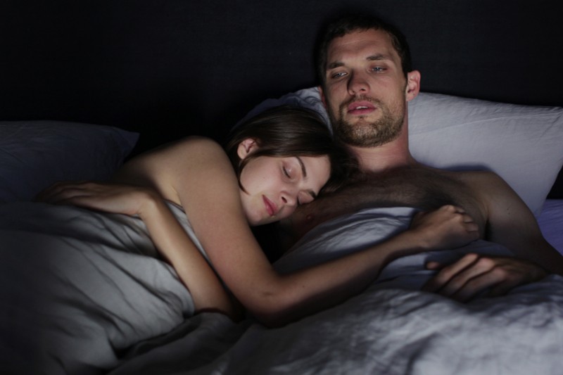Kobieta śpi z głową na piersi mężczyzny, podczas gdy on leży w łóżku, zamyślony i nie może spać. Oboje leżą pod szarymi prześcieradłami i odpoczywają na białych poduszkach w słabo oświetlonym pokoju.
