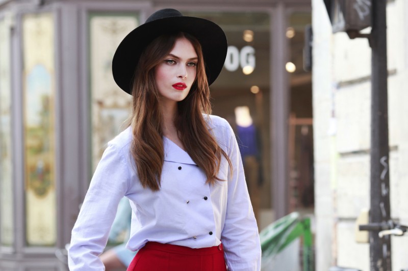 Kobieta o długich brązowych włosach, ubrana w czarny kapelusz z szerokim rondem, białą bluzkę i czerwone spodnie, pewnie stoi na zewnątrz, w miejskim otoczeniu.