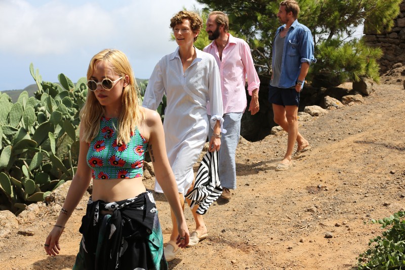 Cztery osoby idą na zewnątrz po ścieżce gruntowej otoczonej kaktusami i zielenią. Kobieta z przodu nosi okulary przeciwsłoneczne i kolorowy krótki top; pozostali idą za nią, ubrani w lekkie, swobodne ubrania. Scena wydaje się słoneczna i zrelaksowana.
