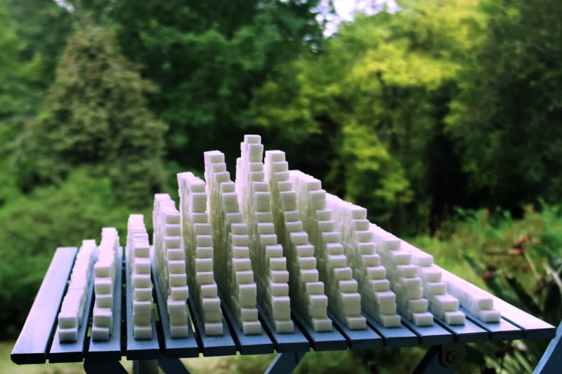 Na blacie stołu leżą starannie ułożone rzędy kostek białego cukru, ułożonych w kształt piramidy o różnej wysokości, na tle bujnej, zielonej, rozmytej zieleni otaczającej teren zewnętrzny.