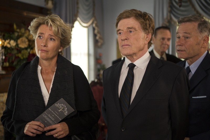 Kobieta i dwóch mężczyzn w formalnych czarnych strojach stoją razem na czymś, co wydaje się być pogrzebem, z poważnymi minami. Kobieta trzyma program, a w tle widać kwiaty.