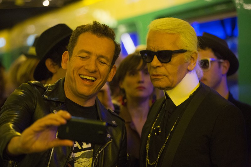 Uśmiechnięty mężczyzna w czarnej skórzanej kurtce robi sobie selfie ze starszym mężczyzną w okularach przeciwsłonecznych i garniturze. Są w pomieszczeniu, z innymi ludźmi i żółto-zielonym tłem za nimi.