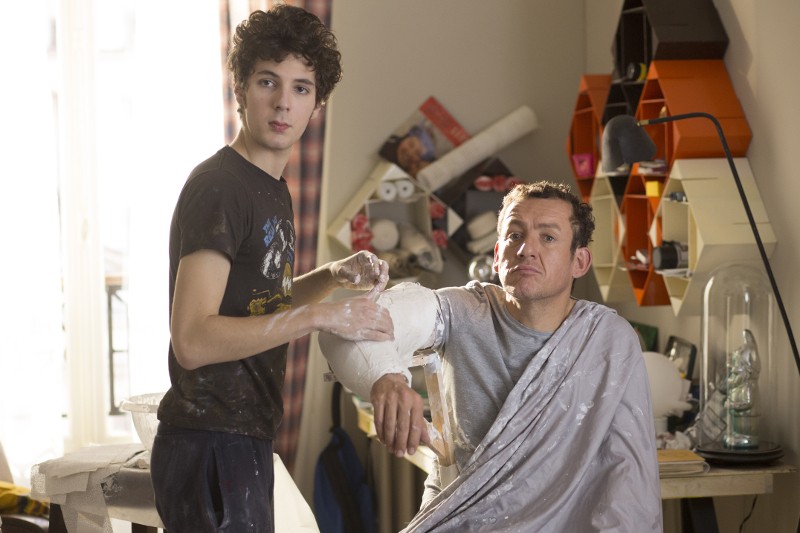 Młody mężczyzna przykrywa ramię starszego mężczyzny gipsem w dobrze oświetlonym, zagraconym pokoju. Starszy mężczyzna siedzi z prześcieradłem na ramionach, podczas gdy młody mężczyzna patrzy w górę, oboje otoczeni przyborami artystycznymi i półkami.