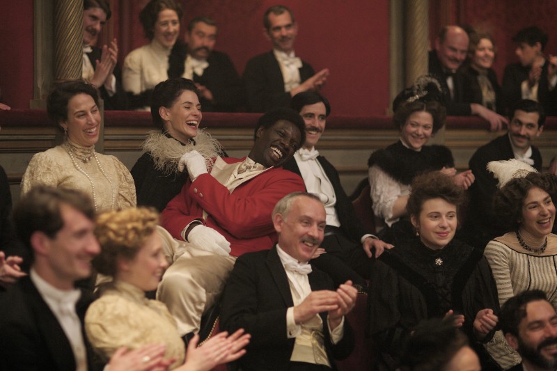 Radosna publiczność teatralna w kostiumach z epoki śmieje się i bije brawo. Mężczyzna w czerwonym płaszczu i kobieta obok niego są szczególnie ożywieni, otoczeni przez innych mężczyzn i kobiety w eleganckich strojach z końca XIX wieku.