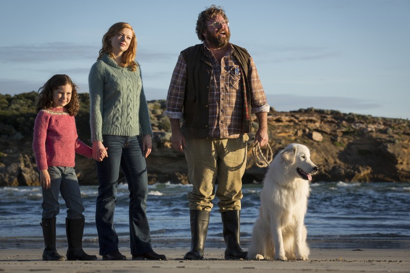 Młoda dziewczyna, kobieta i brodaty mężczyzna z białym psem stoją razem na plaży. Dorośli i dziecko noszą swetry i buty, a mężczyzna trzyma smycz psa, w tle widać fale oceanu i skaliste klify.