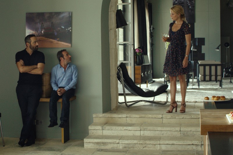 Kobieta w ciemnej, kwiecistej sukience stoi na kamiennych schodach, trzyma kieliszek wina i patrzy na dwóch mężczyzn — jednego stojącego ze skrzyżowanymi ramionami, a drugiego siedzącego na ławce — w nowoczesnym, stylowym salonie.