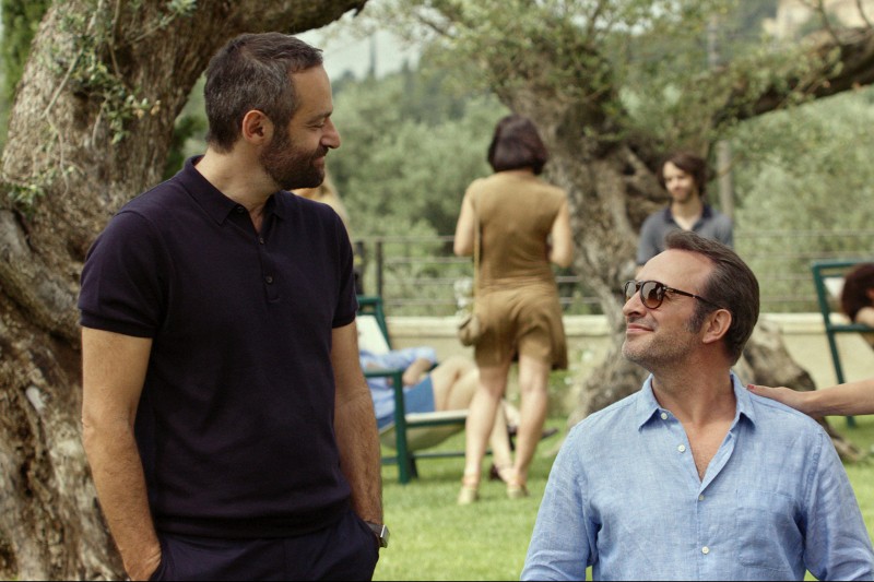 Dwóch mężczyzn stoi na zewnątrz, uśmiechając się do siebie; jeden ma na sobie ciemną koszulkę polo, a drugi jasnoniebieską koszulę z guzikami. Za nimi ludzie odpoczywają na krzesłach ogrodowych pod drzewami w słonecznym otoczeniu ogrodu.