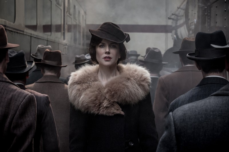 Kobieta w stroju vintage z futrzanym kołnierzem i ciemnym kapeluszem stoi wśród mężczyzn w kapeluszach i płaszczach na zamglonym dworcu kolejowym, patrząc prosto przed siebie. Scena jest nastrojowa i przywołuje scenerię z początku XX wieku.