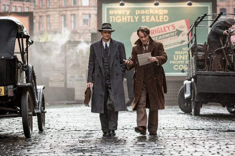 Dwóch mężczyzn w ubraniach w stylu vintage z lat 40. idzie mokrą brukowaną ulicą, jeden trzyma teczkę, a drugi czyta dokument. W tle widać stare samochody i billboard.