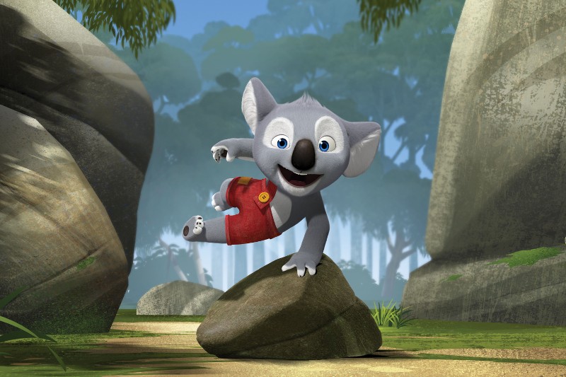 Animowany szary koala ubrany w czerwone spodenki radośnie przeskakuje przez skałę w bujnym, zielonym lesie otoczonym dużymi głazami i drzewami pod jasnym niebem.