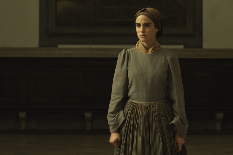 Młoda kobieta w skromnej, staromodnej sukience i chuście na głowie stoi w środku, wyglądając poważnie i napięto, z dłońmi zaciśniętymi po bokach. Tło jest słabo oświetlone ciemnymi drewnianymi panelami.