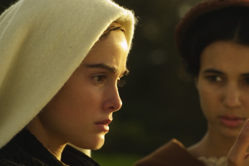 Młoda kobieta z białą chustą na głowie patrzy przed siebie ze łzą na policzku, podczas gdy inna kobieta stoi za nią, lekko nieostra. Obie wydają się poważne i ponure.