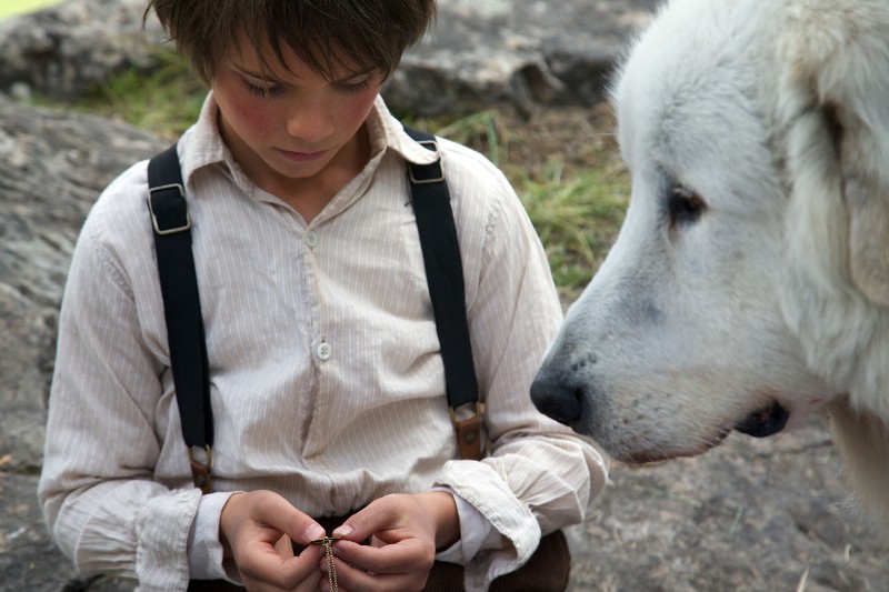Młody chłopiec w białej koszuli z szelkami spogląda w dół na przedmiot w swoich rękach, podczas gdy duży biały pies uważnie go obserwuje. Są na zewnątrz na skalistej powierzchni.