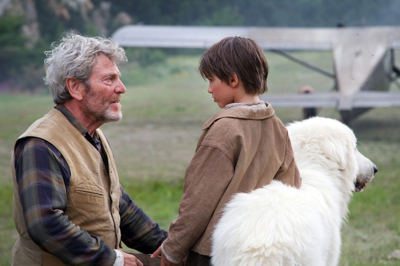 Starszy mężczyzna i chłopiec stoją na zewnątrz, a obok nich jest duży biały pies. Wyglądają, jakby prowadzili poważną rozmowę w pobliżu trawiastego pola, a w tle widać staromodny samolot.