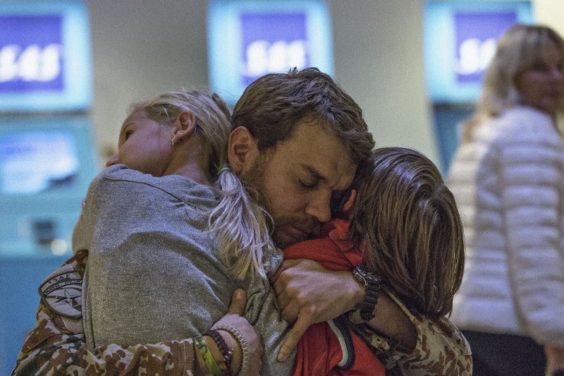 Mężczyzna w mundurze wojskowym mocno obejmuje dwójkę dzieci, z zamkniętymi oczami, wyglądający na wzruszonego. W tle widać rozmazane ekrany odprawy na lotnisku i ludzi, co sugeruje serdeczne spotkanie.