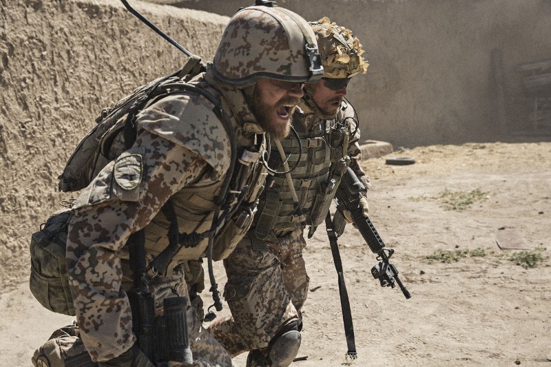 Dwóch żołnierzy w mundurach maskujących i hełmach biegnie obok siebie wzdłuż zakurzonej ściany, uzbrojonych w karabiny i sprzęt taktyczny. Wyglądają na spiętych i skupionych na walce.