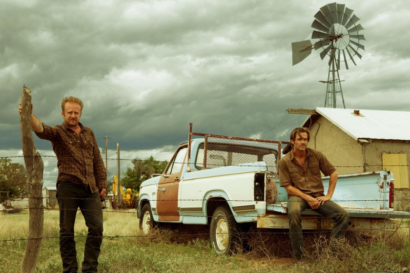 Dwóch mężczyzn w znoszonych ubraniach stoi i siedzi przy starym, białym pickupie na wiejskim polu. W tle pod zachmurzonym niebem widać wiatrak i mały budynek. Scena wygląda na napiętą i zniszczoną.