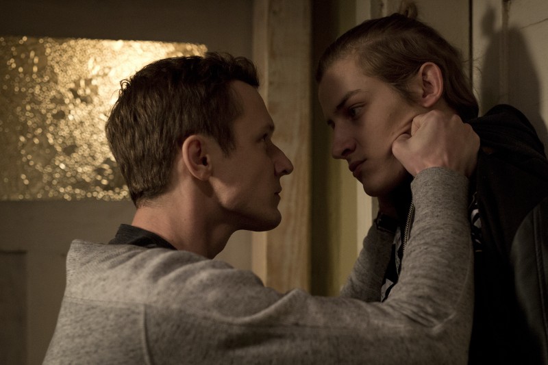 Mężczyzna chwyta innego młodzieńca za kołnierz i przytrzymuje go przy ścianie, uważnie mu się przyglądając. Scena wydaje się napięta, z przyćmionym oświetleniem i poważnymi wyrazami twarzy.