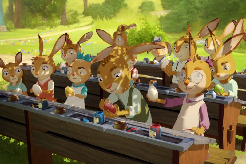 Animowane króliki siedzą przy długich stołach na zewnątrz, malując kolorowe pisanki pędzlami i słoikami z farbą. Scena jest jasna i radosna, osadzona na bujnym zielonym polu ze słońcem i drzewami w tle.