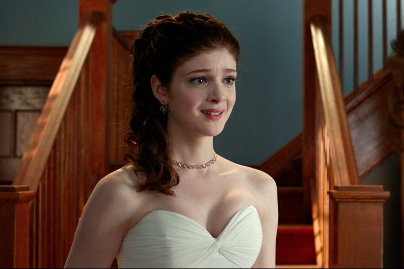 Młoda kobieta z falującymi brązowymi włosami związanymi w kucyk stoi w pomieszczeniu, ubrana w białą sukienkę bez ramiączek i naszyjnik, wygląda na lekko zaniepokojoną lub wzruszoną, stojąc przed drewnianymi schodami.