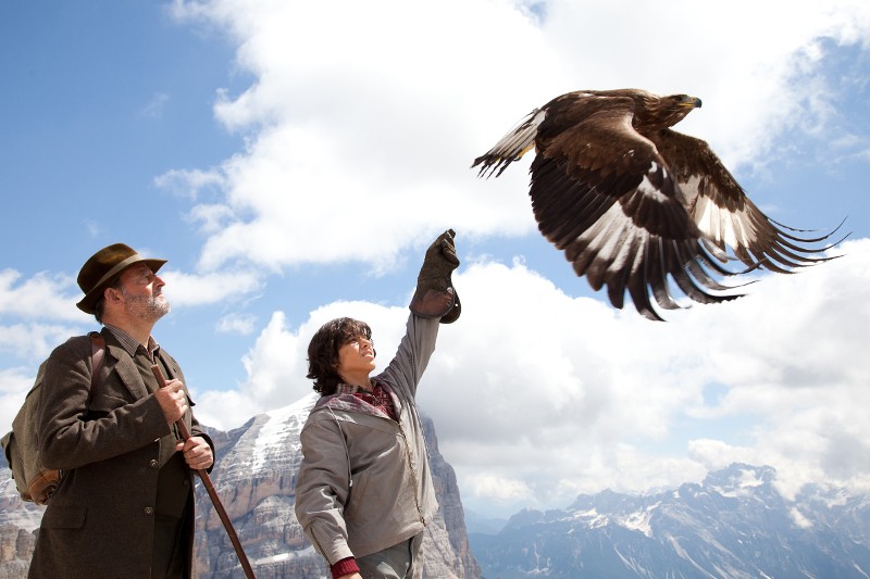 Kobieta w rękawiczce wypuszcza w powietrze dużego orła, a obok niej stoi mężczyzna w kapeluszu. W tle widać ośnieżone góry i częściowo zachmurzone niebo.