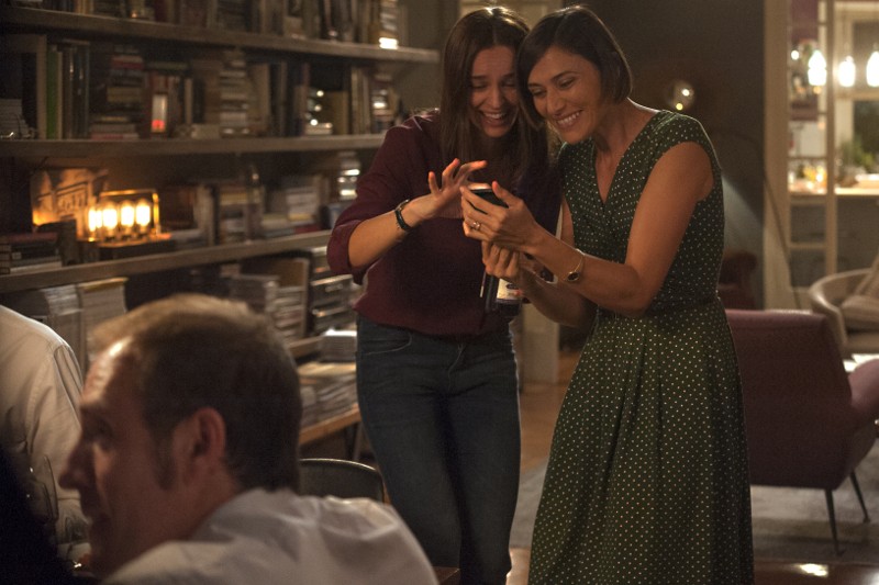 Dwie kobiety uśmiechają się i patrzą razem na smartfon w przytulnym, ciepło oświetlonym pokoju z regałami w tle. Inni ludzie siedzą w pobliżu, tworząc przyjazną, towarzyską atmosferę.