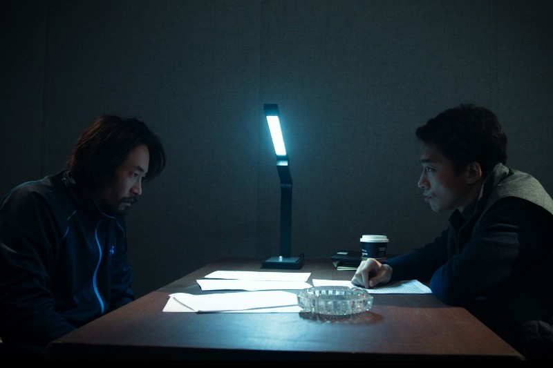 Dwóch mężczyzn siedzi naprzeciwko siebie przy stole w słabo oświetlonym pokoju. Na stole leżą papiery i lampa, co sugeruje intensywną dyskusję lub przesłuchanie. Jeden mężczyzna wygląda poważnie, drugi wydaje się spięty.