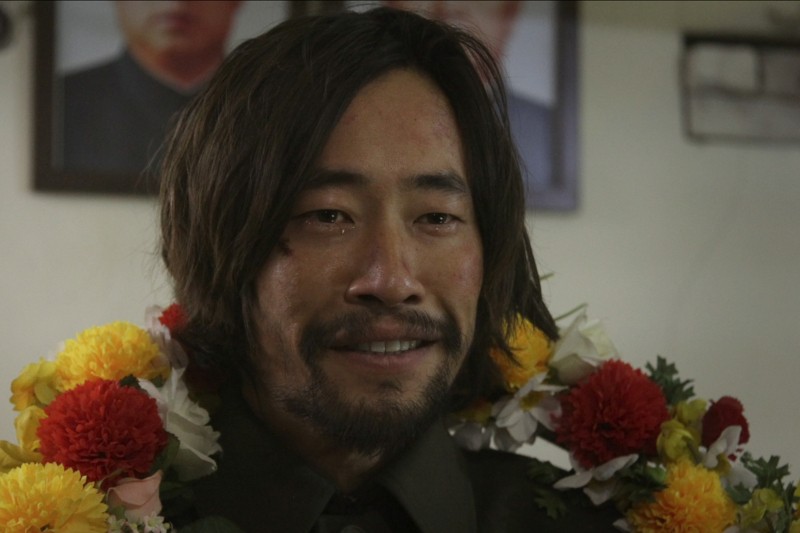 Mężczyzna z włosami do ramion i brodą uśmiecha się łagodnie. Ma na sobie zieloną koszulę i kilka kolorowych girland kwiatów na szyi, a w tle na ścianie wiszą rozmazane portrety.