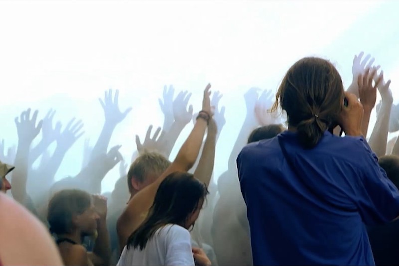 Tłum ludzi z podniesionymi rękami cieszy się ożywionym wydarzeniem na świeżym powietrzu lub koncertem, otoczony mgłą lub dymem. Atmosfera jest energiczna i radosna.